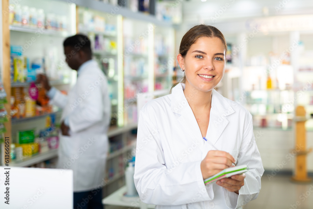 Young smiling female pharmacist working in a pharmacy is standing in the trading floor, making important notes in a notebook