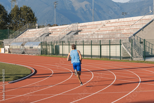 Young man, a professional athlete with a serious and focused face running along an outdoor athletic track near the sports field