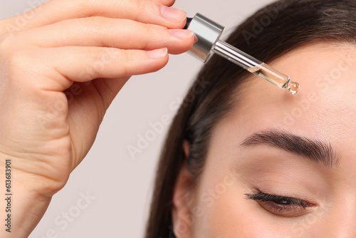 Young woman applying essential oil onto face on light grey background, closeup