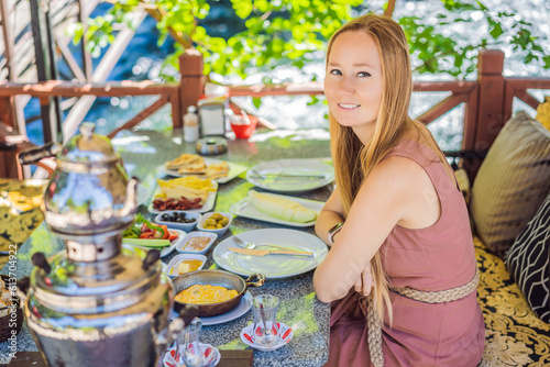 woman tourist eat Turkish breakfast. Turkish breakfast table. Pastries, vegetables, greens, olives, cheeses, fried eggs, spices, jams, honey, tea in copper pot and tulip glasses, wide composition