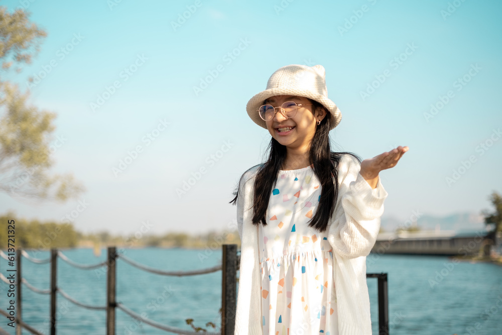 Asian girl in eyeglasses in white dress and hat standing by lake with smiling face in good mood