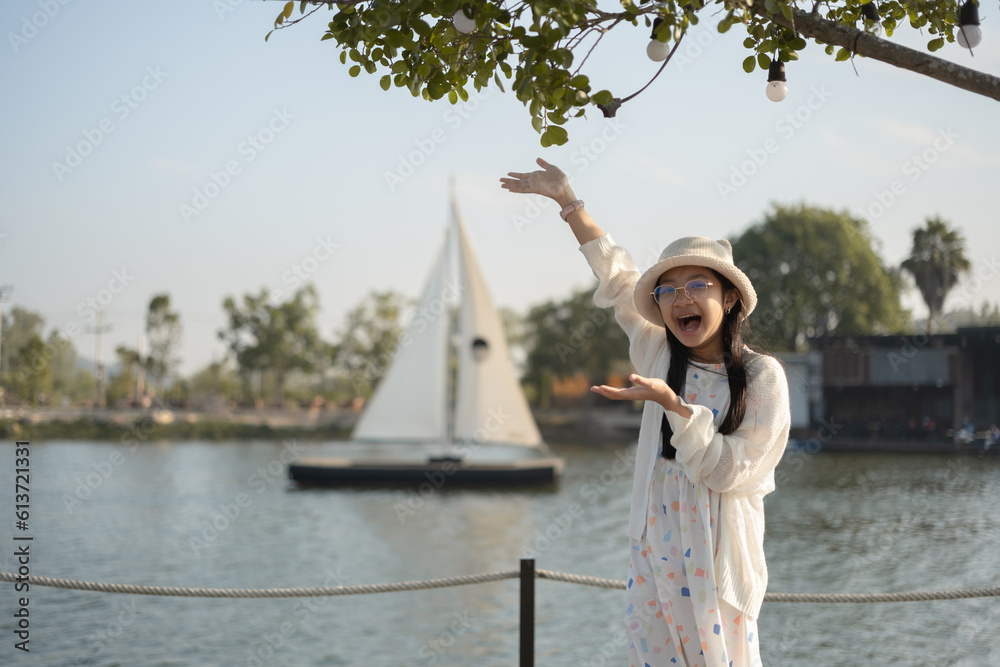 Asian girl in eyeglasses in white dress and hat standing by the lake with a sailboat with a smiling face in a good mood Asian girl in eyeglasses in white dress and hat standing by the lake with a sailboat with a smiling face in a good mood