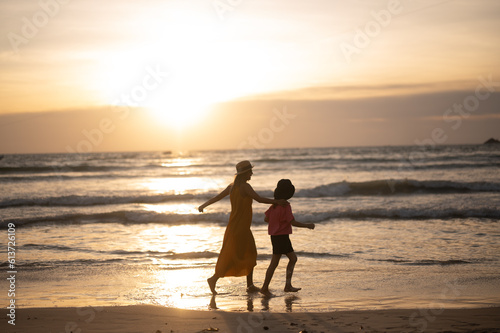 A young mother holds her little daughter by the hand stay holiday together along the beach towards the beautiful silhouette sunset. Happy mom and kid walking a long the white sand beach.
