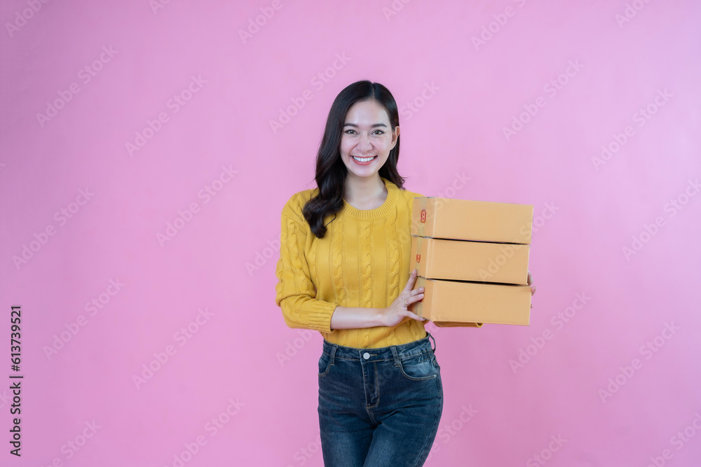 Young Asian woman with long hair, business owner smile powerfully hold 3 parcel boxes Prepare to send orders for SME business products. wearing a yellow coat photo shoot in the pink scene studio Young Asian woman with long hair, business owner smile powerfully hold 3 parcel boxes Prepare to send orders for SME business products. wearing a yellow coat photo shoot in the pink scene studio