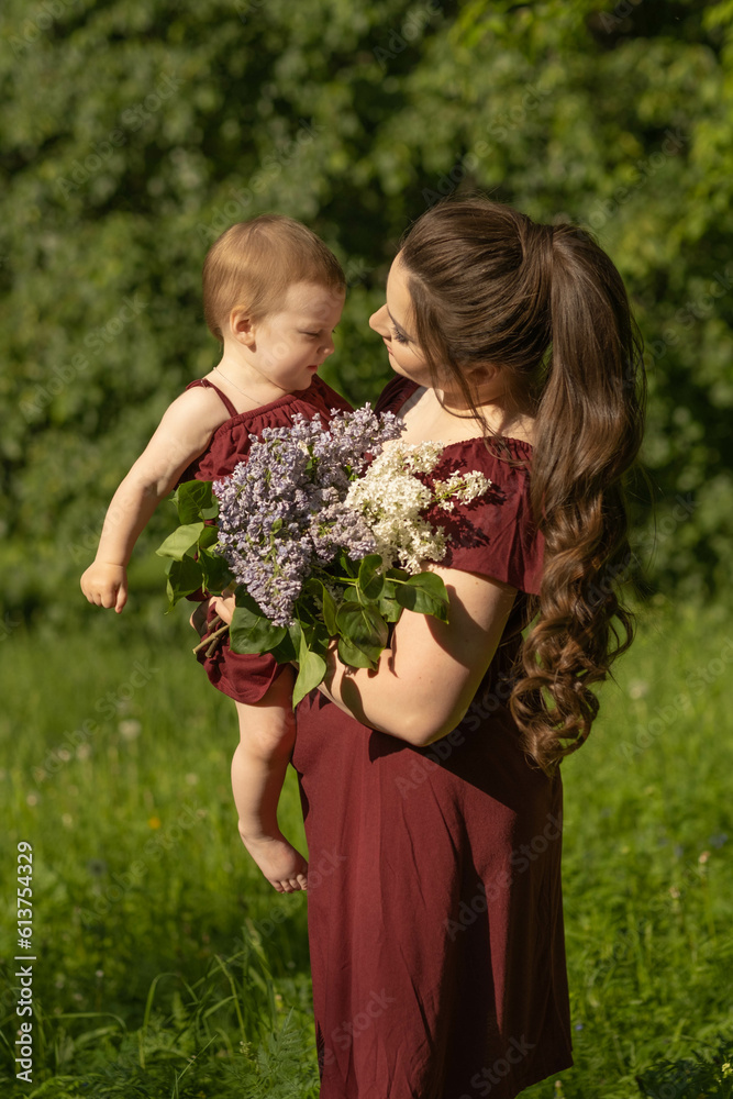 Mother's Day celebration outdoors, a happy mother carrying her one-year-old baby girl and a bunch of lilacs on a clear, sunny day Mother's Day celebration outdoors, a happy mother carrying her one-year-old baby girl and a bunch of lilacs on a clear, sunny day