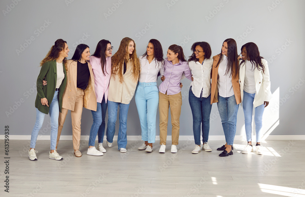 Full size group of a cheerfull happy young diverse women friends or coworkers and company employees looking at each other hugging and smiling together standing on a grey wall background.