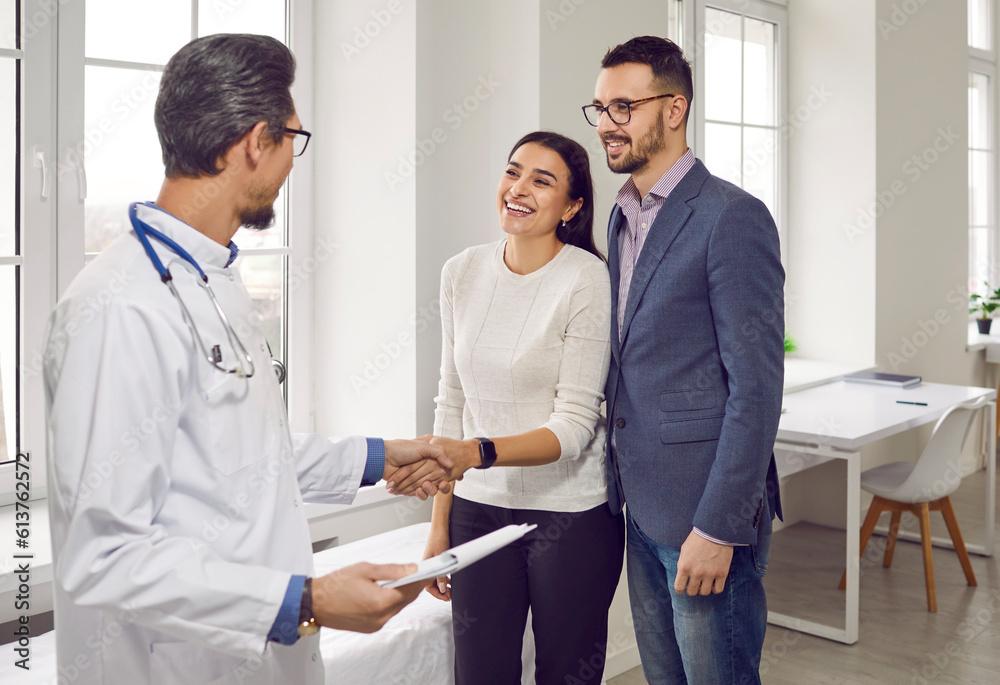 Fototapeta premium Young beautiful couple at doctor's consultation in the office of the Family planning medical center. Professional obstetrician-gynecologist talks to happy family, shaking hands with smiling girl.