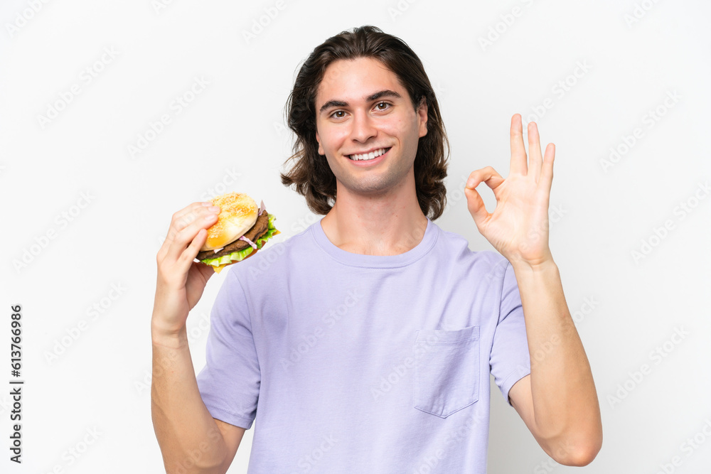 Young handsome man holding a burger isolated on white background showing ok sign with fingers Young handsome man holding a burger isolated on white background showing ok sign with fingers