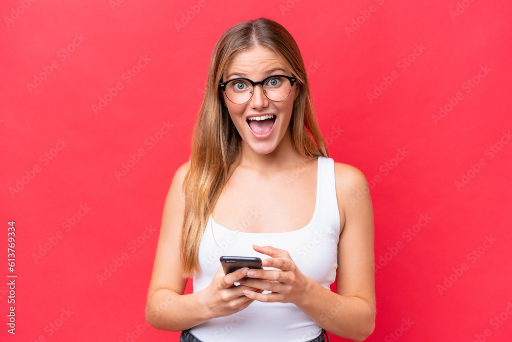 Young beautiful woman isolated on red background surprised and sending a message Young beautiful woman isolated on red background surprised and sending a message