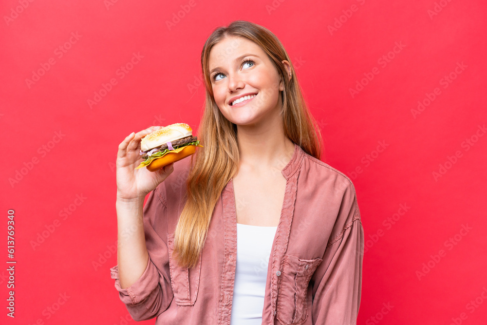Young caucasian woman holding a burger isolated on red background looking up while smiling Young caucasian woman holding a burger isolated on red background looking up while smiling
