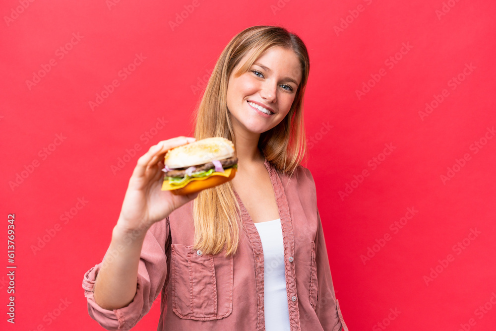 Young caucasian woman holding a burger isolated on red background with happy expression Young caucasian woman holding a burger isolated on red background with happy expression