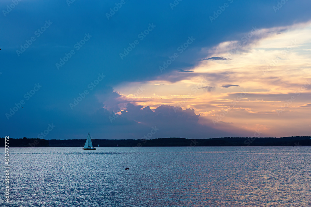 Beautiful landscape of water, little yacht on horizon, sky at sunset before storm: blue huge cloud begins to cover colorful sky; natural picturesque evening scenery of river or lake, summer background