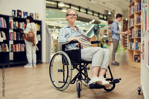 Portrait of happy smart person with disability girl with blond hair sitting in wheelchair and holding books in university library