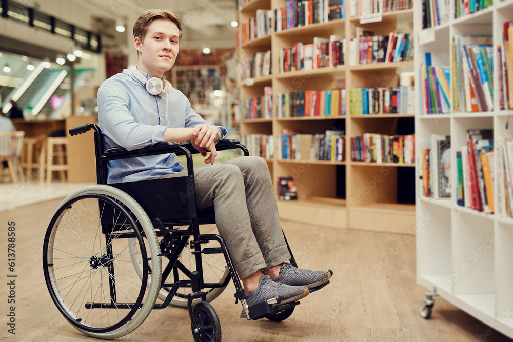 Portrait of content student boy with paralyzed legs sitting in wheelchair and choosing books in library Portrait of content student boy with paralyzed legs sitting in wheelchair and choosing books in library