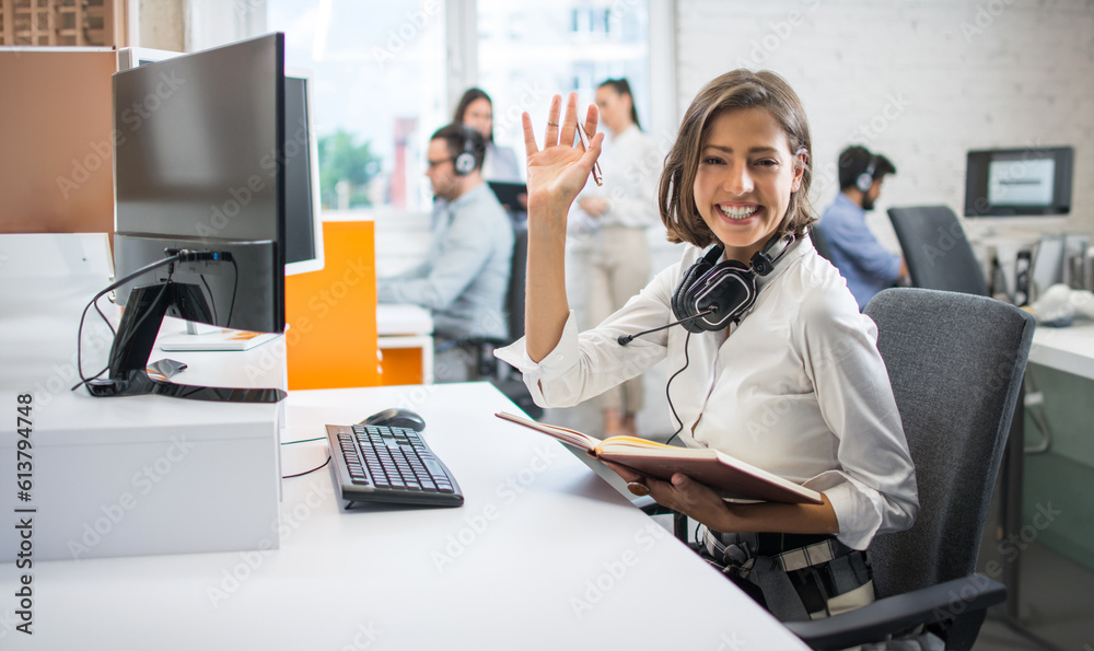 Obraz premium Portrait of friendly businesswoman holding pen and notebook and waving hand to camera at office.