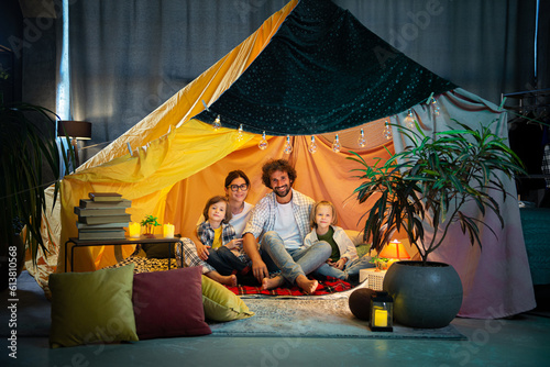 Big young family mother dad and two kids speeding time together while watching a movie on the laptop and eating some popcorn at home in a large handmade tent