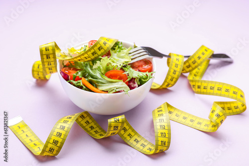 Bowl with fresh vegetable salad  fork and measuring tape on violet background. Healthy diet concept