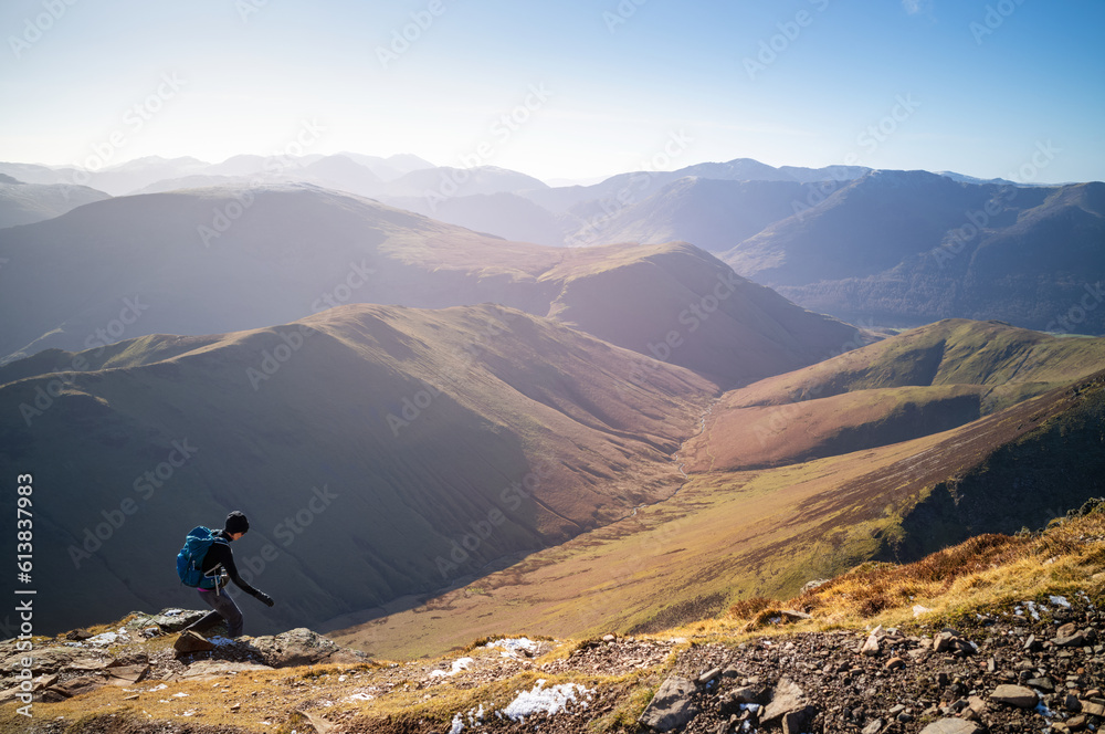 A female hiker descending a rocky path towards Sail from Crag Hill with distant views of High Stile and Red Pike.