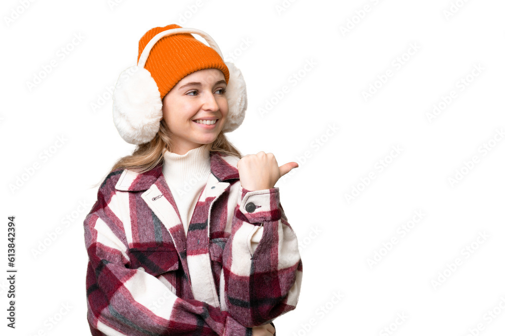 Young English woman wearing winter muffs over isolated background pointing to the side to present a product Young English woman wearing winter muffs over isolated background pointing to the side to present a product