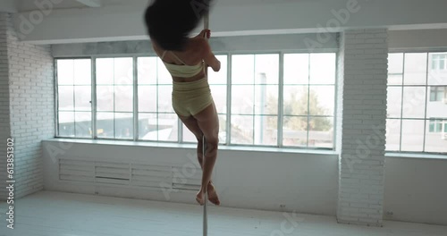Asian strip dancer spinning on a pylon, in a bright white studio in daylight against the background of a panoramic window in beige lingerie. Sexual sensual graceful movements of young woman