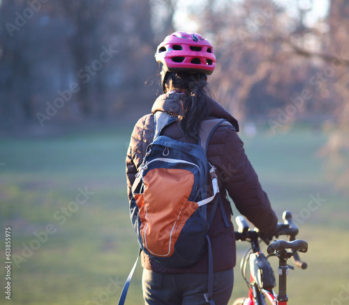 woman in helmet wearing jacket,backpack holding bicycle looking away from camera (no face) at sunset soft golden hour light (nature, park background with trees and grass) fall, winter, spring bike
