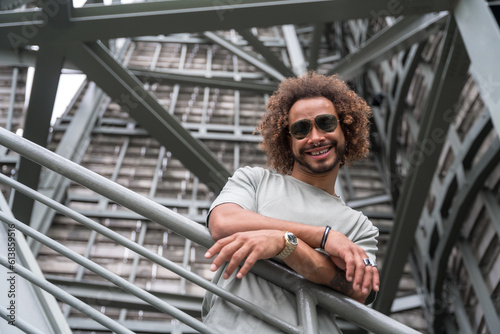 Portrait of a young man with afro hair wearing sunglasses on the stairs in the city smiling, urban fashion cool concept of a hipster guy with stylish