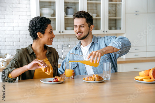 Happy young spouses  an african american woman and a caucasian man  have breakfast together at home in the kitchen  eat croissants and drink juice  communicate  look at each other  smile