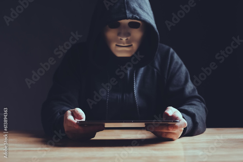 Black hat wearing white mask hacker in hood using tablet on desk to hacking privacy sensitive data cyber crime hack in dark room background. Cyber security cyber crime concept. Hacking phishing