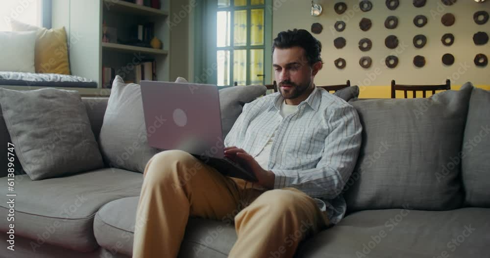 A young man in casual clothes smiles while typing on a laptop while sitting on the couch at home