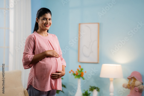 Happy smiling Indian Pregnant woman standing by feeling tummy while looking camera at home - concept of motherhood, expecting baby and parenthood