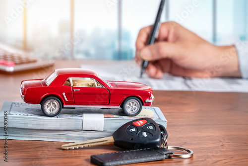 Car loan or buying a new car concept. Red car on dollar banknotes and car key on table with a man signing on paper in background.