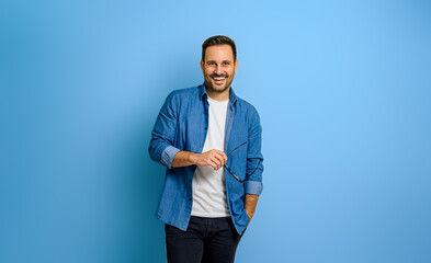 Happy handsome businessman with hand in pocket holding eyeglasses and standing over blue background