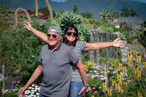 Cheerful senior couple  in denim and sunglasses enjoying a sunny day in beautiful garden with waterfall in the background. Free happy retirement concept