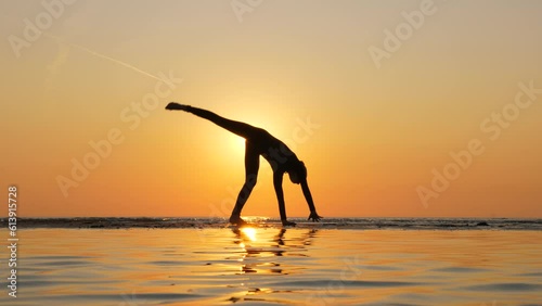 Skilled and graceful acrobat, slender girl effortlessly performs cartwheel against backdrop of vibrant sunset. Bright sun shining behind her creates stunning silhouetted appearance