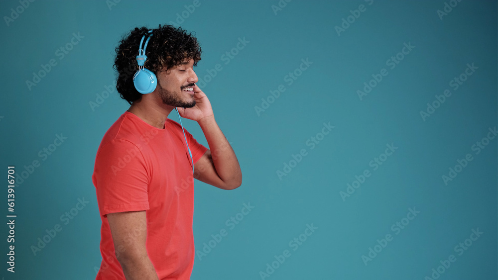 Young smiling happy cheerful student swarthy man dancing with headphones listen to music have fun in coral t-shirt on blue studio background. People lifestyle concept Young smiling happy cheerful student swarthy man dancing with headphones listen to music have fun in coral t-shirt on blue studio background. People lifestyle concept