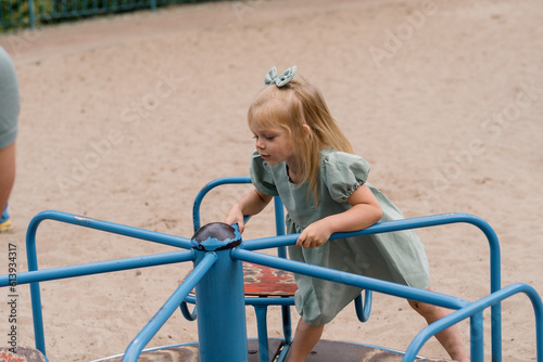 Little cute upset girl in a green summer dress with a bow on her head on the playground rides on the swing alone children without adult supervision