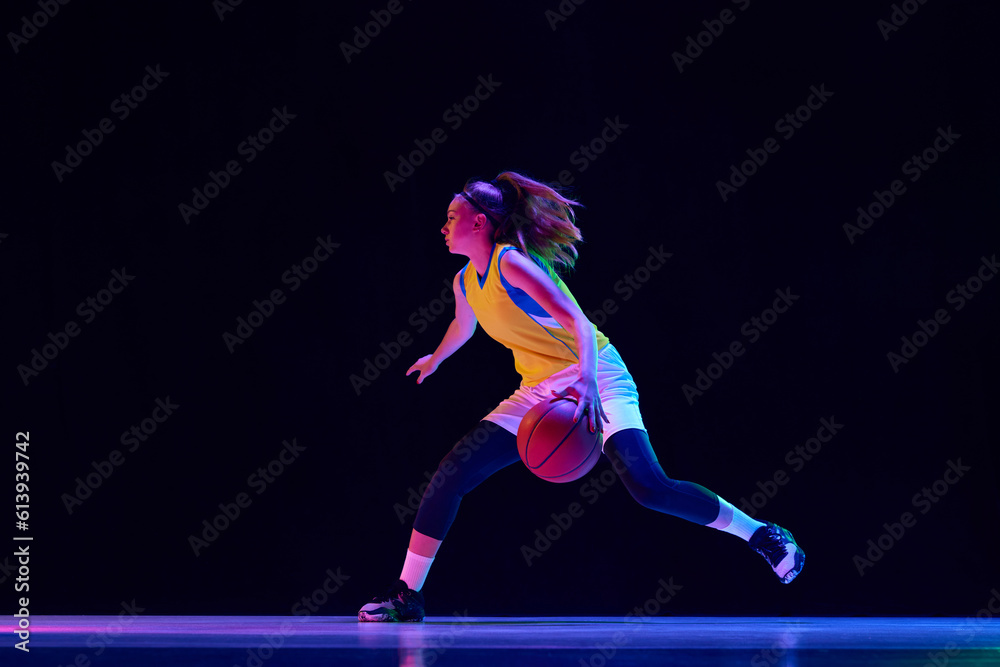 Young female athlete, professional basketball player in motion, dribbling ball over black studio background in neon light. Professional sport, action and motion, game, competition, hobby, ad concept