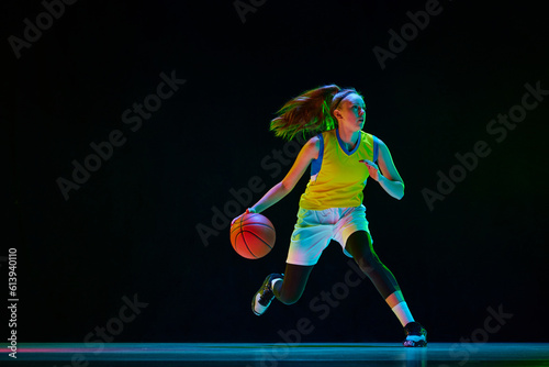 Young motivated girl, basketball player in uniform running with ball against black studio background in neon light. Concept of professional sport, action and motion, game, competition, hobby, ad © master1305