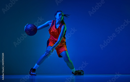 Dynamic image of young girl  basketball player in uniform in motion  playing over blue studio background in neon light. Concept of professional sport  action and motion  game  competition  hobby  ad