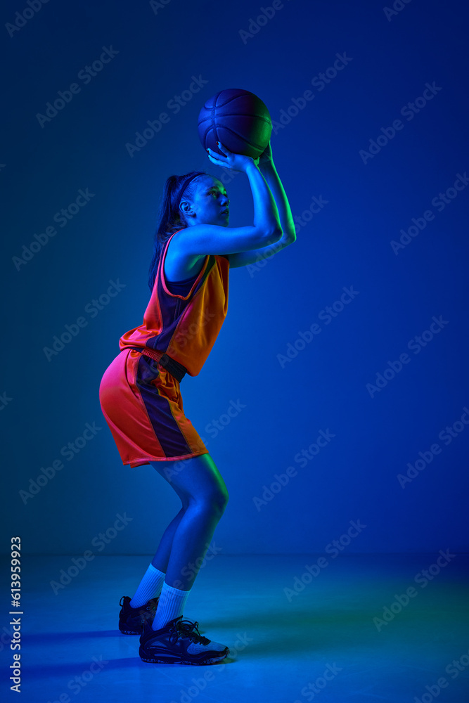 Winning goal. Side view of concentrated female basketball player throwing ball against blue studio background in neon light. Professional sport, action and motion, game, competition, hobby, ad concept