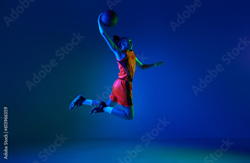 Slam dunk. Motivated young girl, basketball player throwing ball in jump against blue studio background in neon light. Concept of professional sport, action and motion, game, competition, hobby, ad © master1305