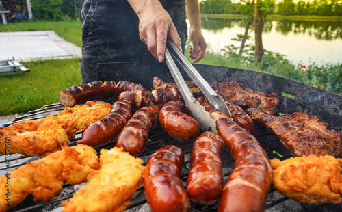 Man turning the sausage on the bbq with kitchen tongs. Person grilling in the garden. Barbeque with sausages and fried fish on the grill. 