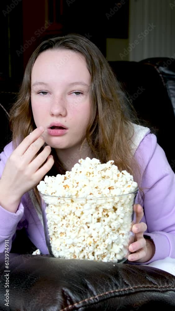 Watching a movie with eyes wide open eating popcorn open wide-eyed teenage girl sitting in dark room on the couch watching TV with her mouth eating flakes of delicious Watching a movie with eyes wide open eating popcorn open wide-eyed teenage girl sitting in dark room on the couch watching TV with her mouth eating flakes of delicious
