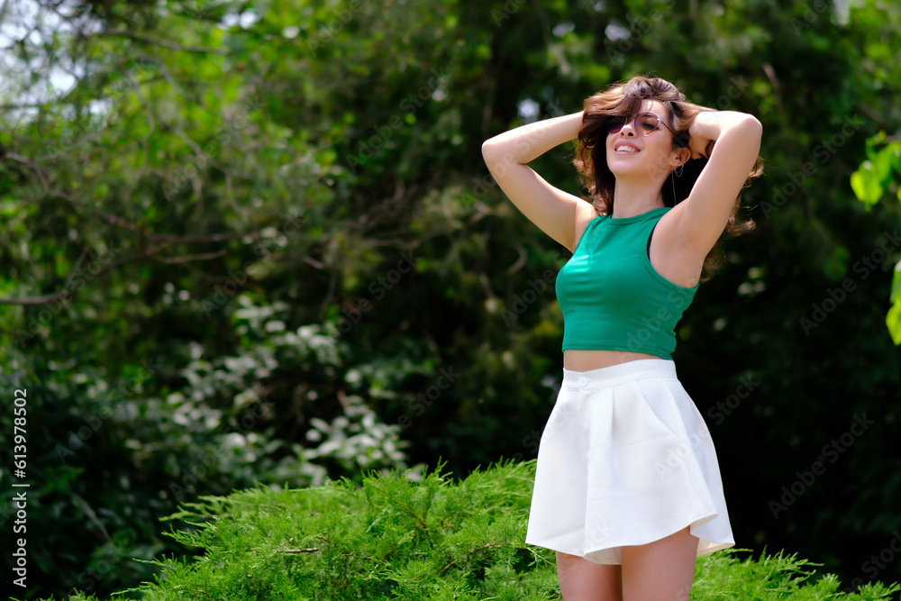 Beautiful girl in a green top and white shorts in a city park on a sunny summer day. Posing with hands holding hair. laughing Beautiful girl in a green top and white shorts in a city park on a sunny summer day. Posing with hands holding hair. laughing