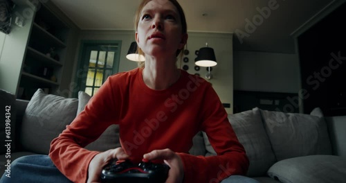 A young woman in everyday ojeda plays video games using a joystick while sitting on the couch at home. The video moves from the carpet to the woman's face photo
