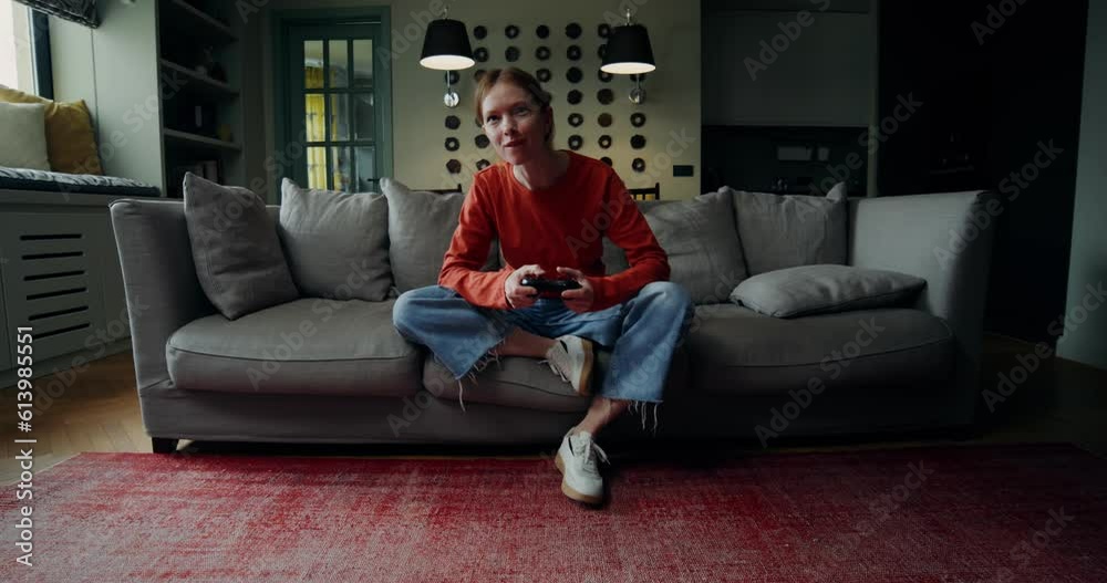 A young woman in casual clothes is smiling while playing video games with a joystick, sitting on the couch at home. The video moves from the general plan to the woman's face A young woman in casual clothes is smiling while playing video games with a joystick, sitting on the couch at home. The video moves from the general plan to the woman's face