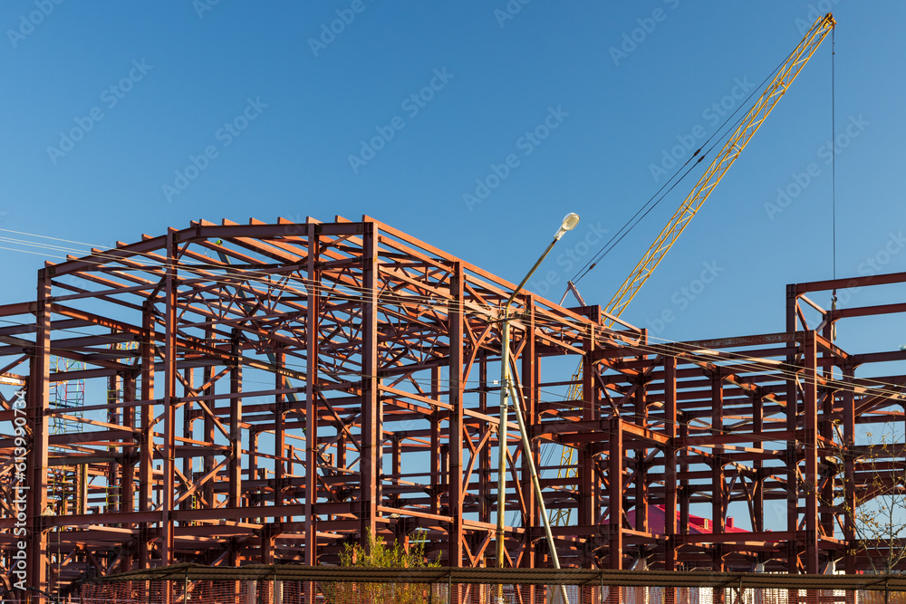 View of the construction site. The metal frame of a building under construction and a crane against a cloudless blue sky. Construction of the school building.