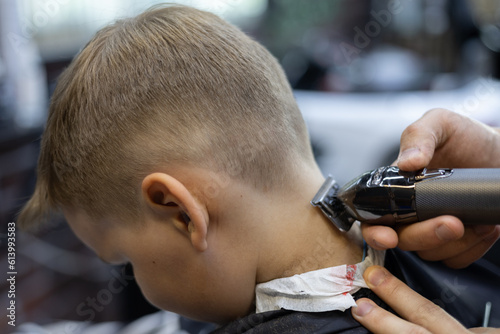 The barber's hand shaves the hair on the back of the child's head in a beauty salon The child is given a light haircut for the summer Child boy is not afraid to go to the hairdresser to do a haircut.