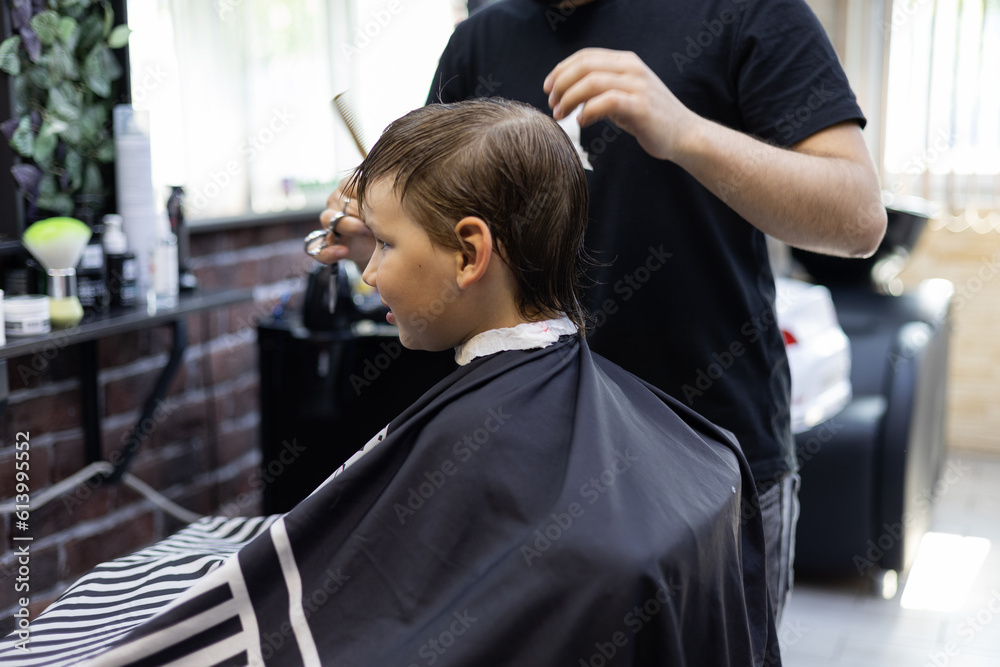 Child boy sitting in a chair in a barbershop carefully watching the work of a hairdresser in the mirror. The hairdresser's hands deftly and quickly cut off the regrown hair of a child. Child boy sitting in a chair in a barbershop carefully watching the work of a hairdresser in the mirror. The hairdresser's hands deftly and quickly cut off the regrown hair of a child.