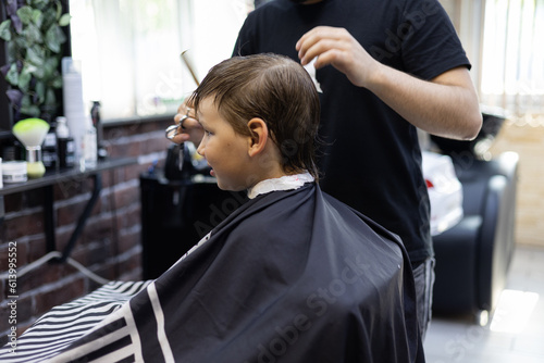 Child boy sitting in a chair in a barbershop carefully watching the work of a hairdresser in the mirror. The hairdresser's hands deftly and quickly cut off the regrown hair of a child.